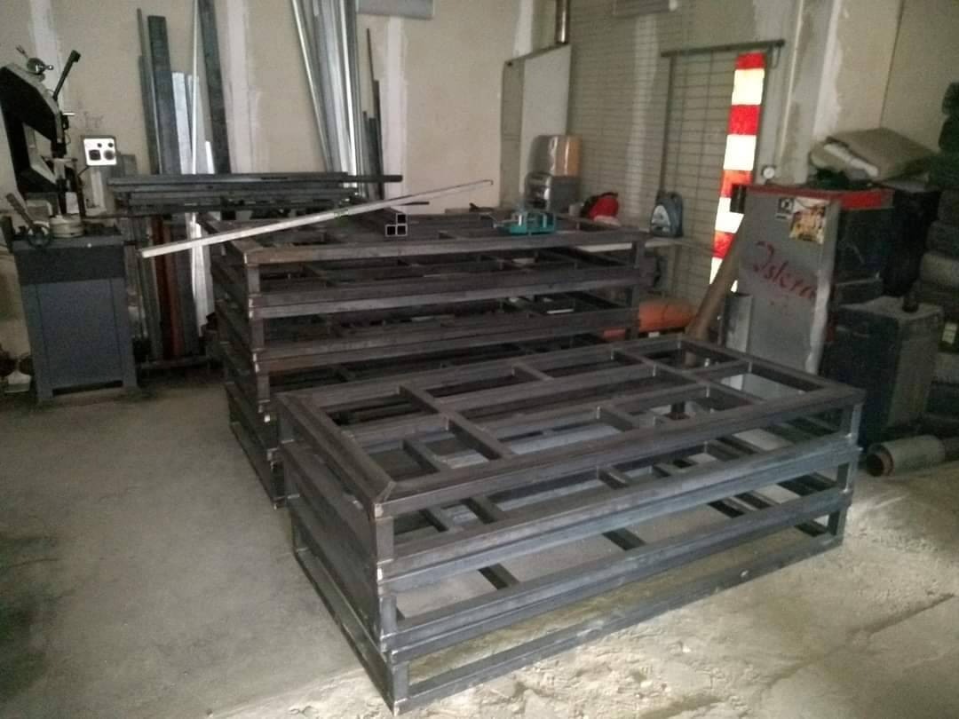 Stos metalowych, spawanych ram o prostokątnych oczkach, ułożonych w warsztacie, widoczna piła taśmowa do metalu i butla z gazem.