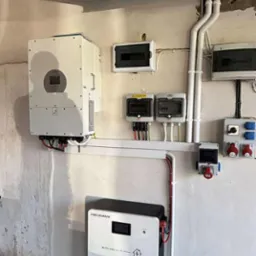 Kompletna instalacja fotowoltaiczna w garażu: inwerter, magazyn energii Heckman, rozdzielnice, kable i gniazda. Białe urządzenia na tle surowej ściany.