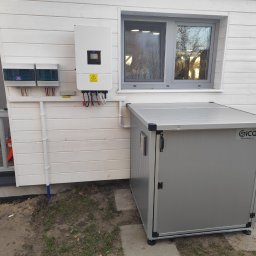 GLOBCORE Sp. z o.o. - Biała skrzynka z instalacją elektryczną i szarym magazynem na zewnątrz domu z drewnianą elewacją. Widoczne okno i elementy instalacji.