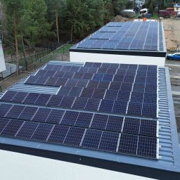 GLOBCORE Sp. z o.o. - Dwa budynki z panelami słonecznymi na dachu, widok z góry. Na drugim planie las i zabudowa mieszkalna. Na dachu jednego z budynków brakuje kilku paneli.