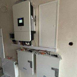 GLOBCORE Sp. z o.o. - Zamontowany inwerter i dwa akumulatory Deye na ścianie wewnątrz budynku. Widoczne okablowanie i elementy instalacji. Biała ściana z widocznymi niedoskonałościami.