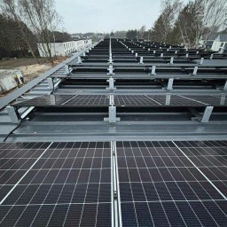 GLOBCORE Sp. z o.o. - Rząd paneli fotowoltaicznych na szarym dachu budynku, zamontowanych na metalowych wspornikach. W tle drzewa i budynki w pochmurny dzień.