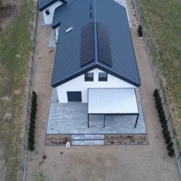 GLOBCORE SPÓŁKA Z OGRANICZONĄ ODPOWIEDZIALNOŚCIĄ - Widok z drona na dom z ciemną dachówką i panelami słonecznymi. Jasna elewacja, szara kostka brukowa na tarasie i zadbany ogród z młodymi tujami wzdłuż ogrodzenia.