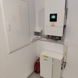 GLOBCORE SPÓŁKA Z OGRANICZONĄ ODPOWIEDZIALNOŚCIĄ - Biała instalacja z inwerterem i akumulatorem Daye Solar na ścianie, obok skrzynka elektryczna i gaśnica. Widoczne kable i oznaczenia ostrzegawcze.