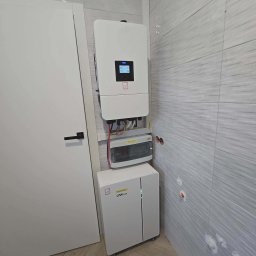 GLOBCORE SPÓŁKA Z OGRANICZONĄ ODPOWIEDZIALNOŚCIĄ - Biały inwerter fotowoltaiczny Deye i magazyn energii DAH Solar zamontowane na ścianie z szarymi płytkami wewnątrz pomieszczenia, obok białe drzwi.