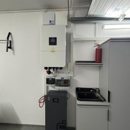GLOBCORE SPÓŁKA Z OGRANICZONĄ ODPOWIEDZIALNOŚCIĄ - Zamontowany, kompletny system zasilania awaryjnego w garażu: biały inwerter, szary akumulator, skrzynka bezpiecznikowa, okablowanie. Gaśnica na półce.