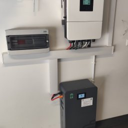 GLOBCORE Fotowoltaika - Zamontowany inwerter i akumulator energii na ścianie, połączone przewodami. Widoczna skrzynka rozdzielcza i fragment podłogi z zabawką. Gotowa instalacja.