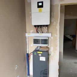 GLOBCORE Fotowoltaika - Szary akumulator energii na kółkach i biały inwerter na ścianie w surowym wnętrzu. Widoczne kable i instalacja elektryczna. Etap montażu.