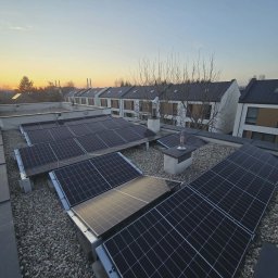 GLOBCORE Fotowoltaika - System paneli słonecznych na płaskim dachu pokrytym jasnym żwirem, z widocznymi kominami, na tle nowoczesnych domów szeregowych podczas zachodu słońca.