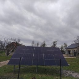 GLOBCORE Fotowoltaika - Naziemna instalacja paneli słonecznych na metalowej konstrukcji, na trawiastym terenie z siatkowym ogrodzeniem. W tle wiejskie zabudowania i bezlistne drzewa pod pochmurnym niebem.