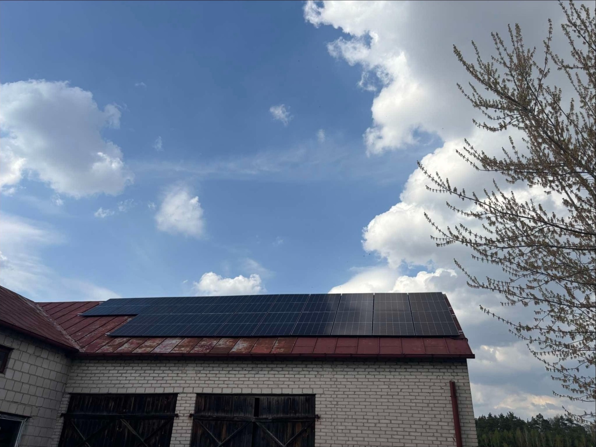 Panele słoneczne na dachu budynku gospodarczego z białej cegły, na tle błękitnego nieba z chmurami i drzewa z młodymi liśćmi.