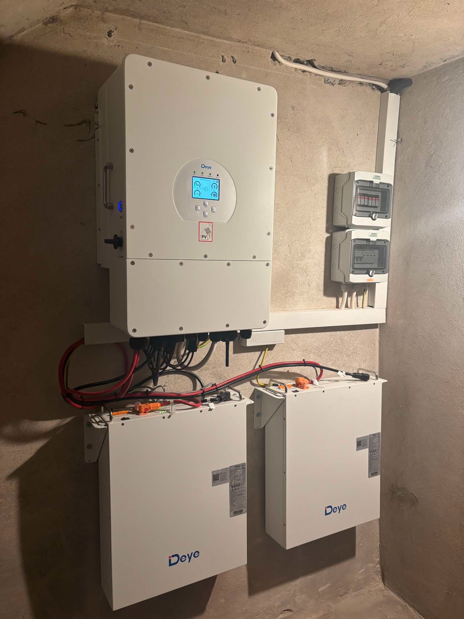 Zamontowany system magazynowania energii z dwoma akumulatorami i inwerterem na ścianie w surowym wnętrzu, widoczne przewody oraz zabezpieczenia elektryczne.