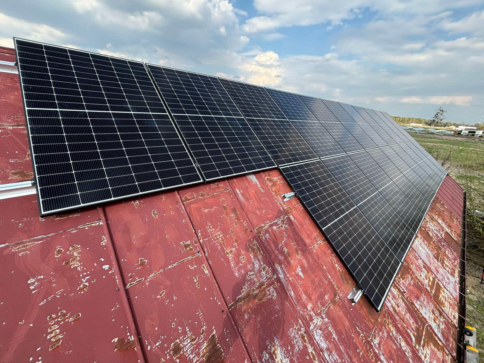 Panele fotowoltaiczne zamontowane na zardzewiałym, czerwonym dachu z blachy trapezowej, widok na pole i błękitne niebo z chmurami.