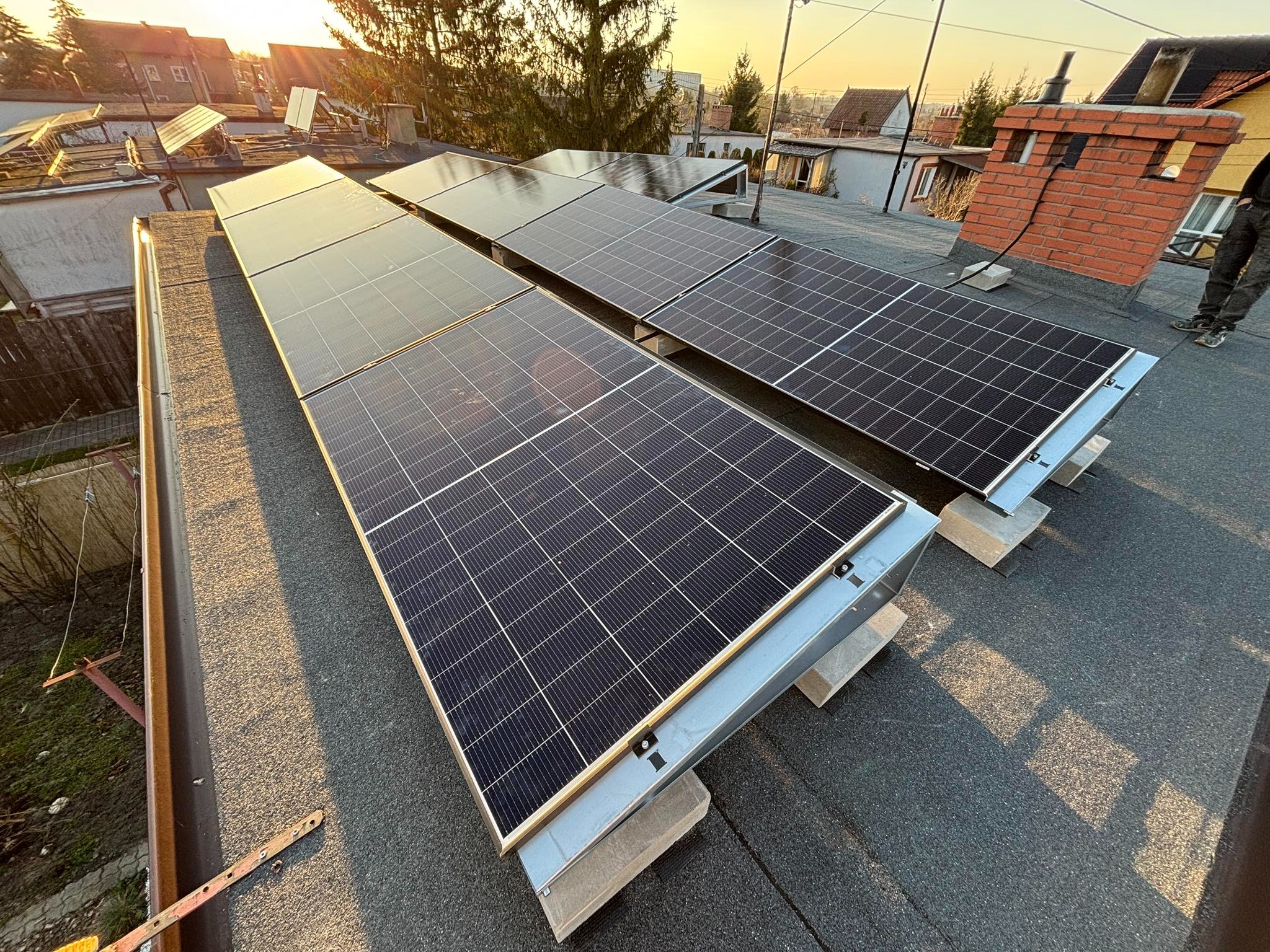 Panele słoneczne na płaskim dachu w Żyrardowie, zamontowane na betonowych wspornikach, widok z boku z fragmentem komina w tle. Częściowo widoczna sylwetka instalatora.