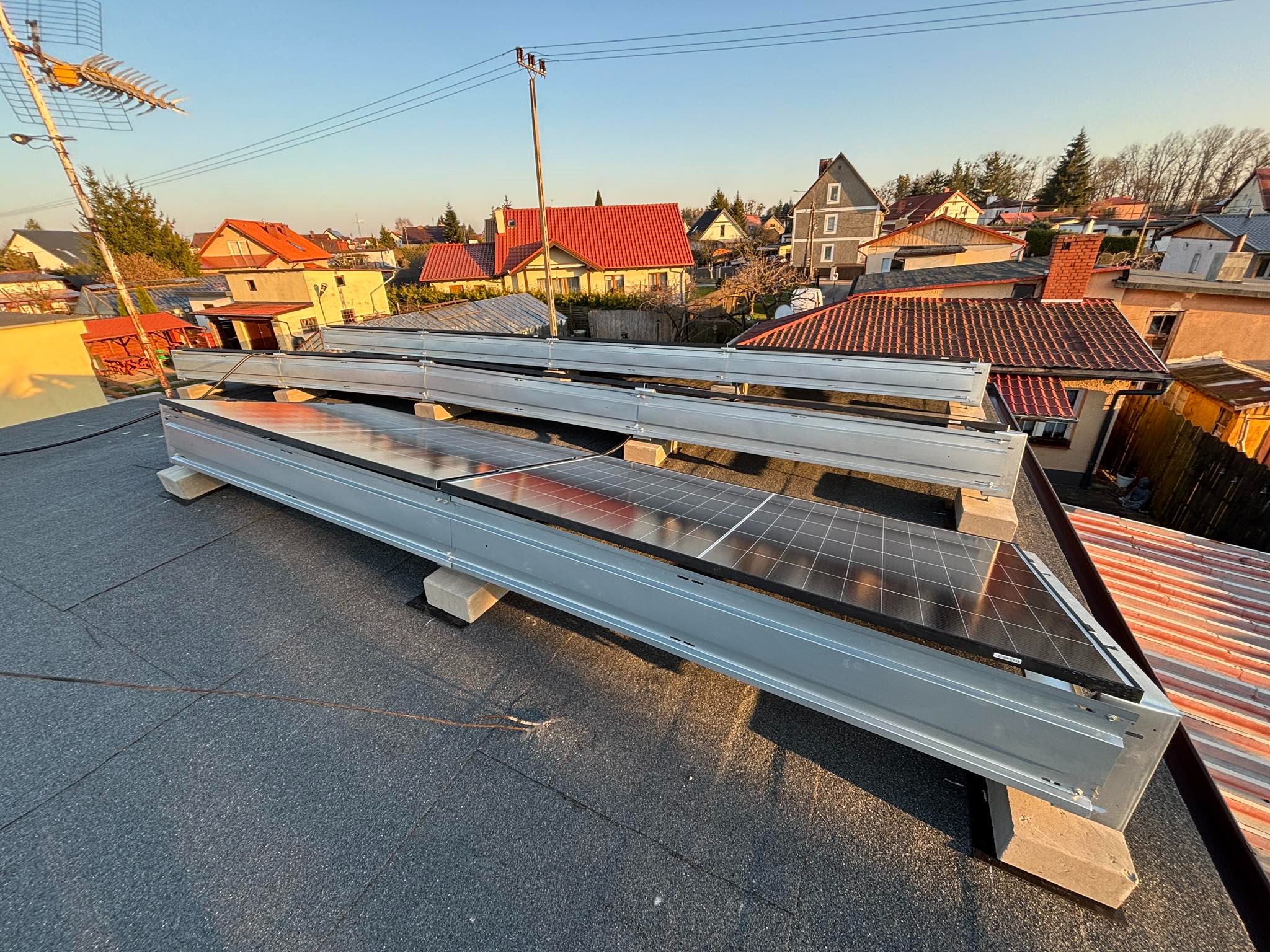 Montaż paneli słonecznych na płaskim dachu z widocznymi elementami konstrukcyjnymi i betonowymi wspornikami, w tle zabudowa jednorodzinna oświetlona popołudniowym słońcem.