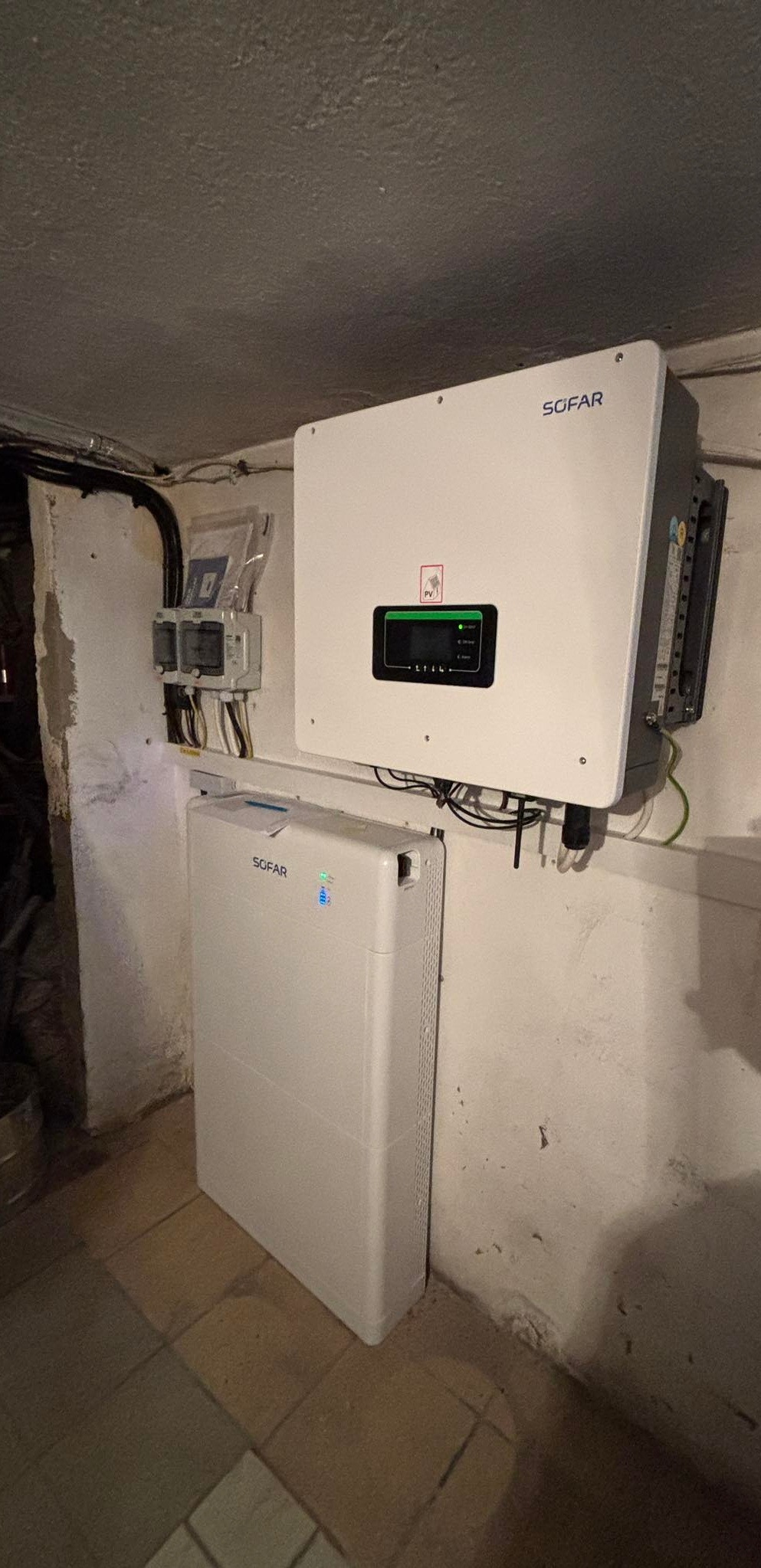Biały magazyn energii i falownik marki SOFAR zamontowane na ścianie w pomieszczeniu technicznym, widoczne również liczniki energii.