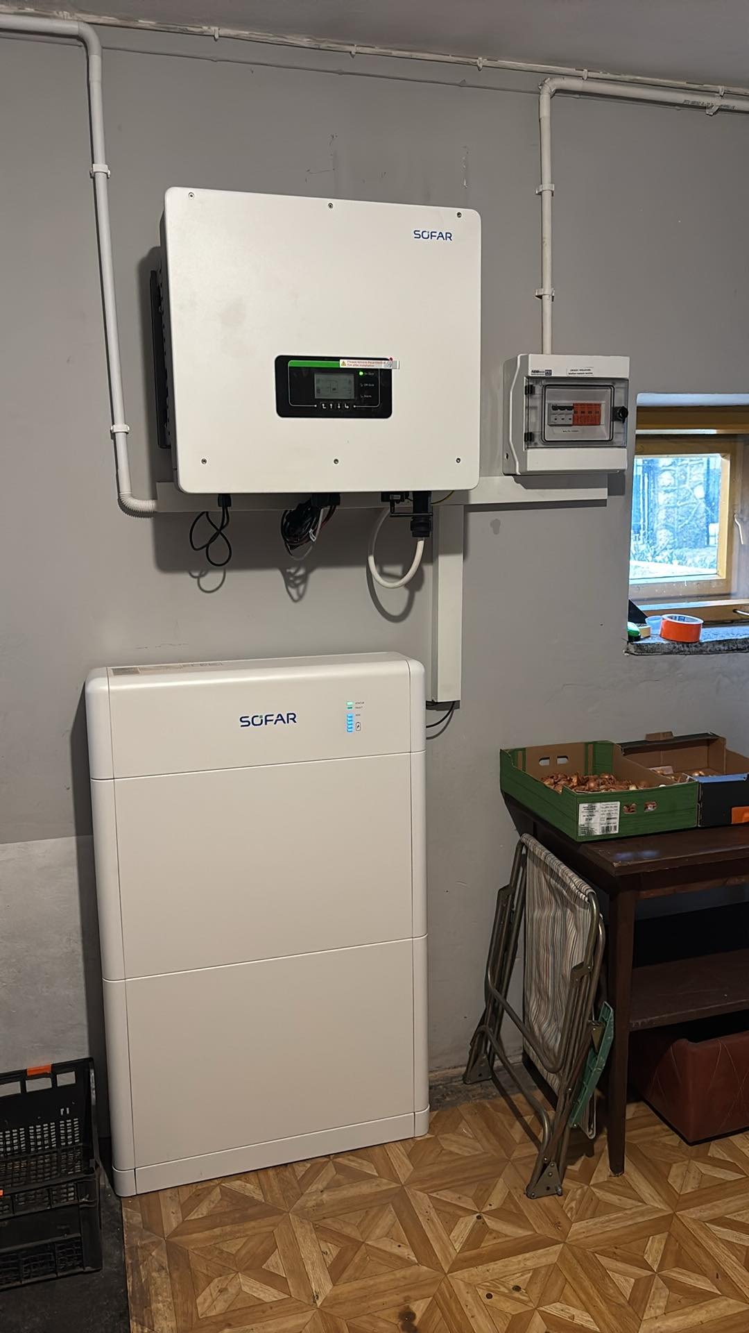 Biały magazyn energii SOFAR zamontowany na szarej ścianie, obok skrzynka elektryczna i okno. Na podłodze wzorzysty parkiet, obok stolik z kartonami cebuli i składane krzesło.