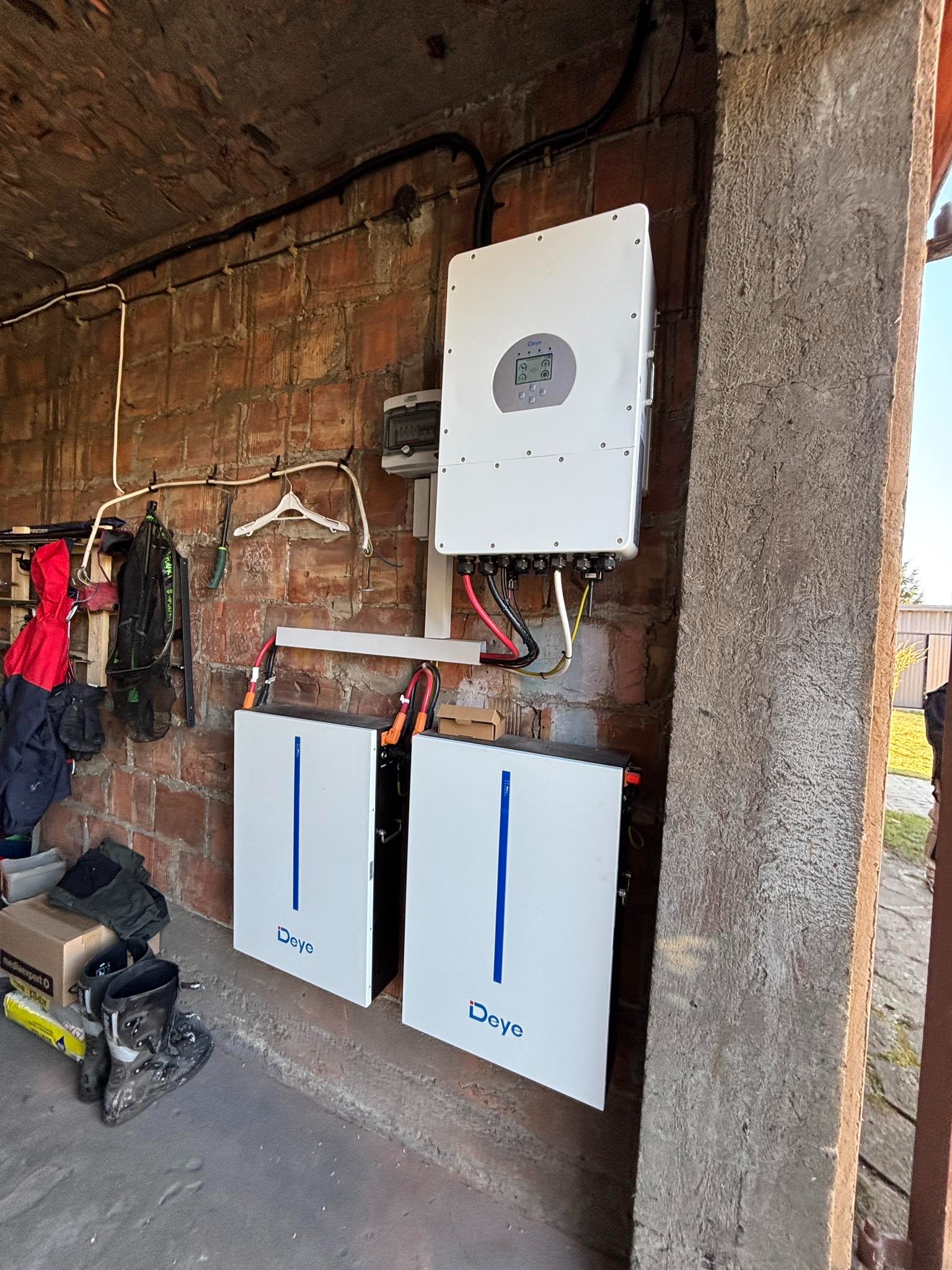 Dwa białe magazyny energii Deye z niebieskimi paskami, podłączone do białego inwertera na ceglanej ścianie w garażu, obok wiszą ubrania i narzędzia.
