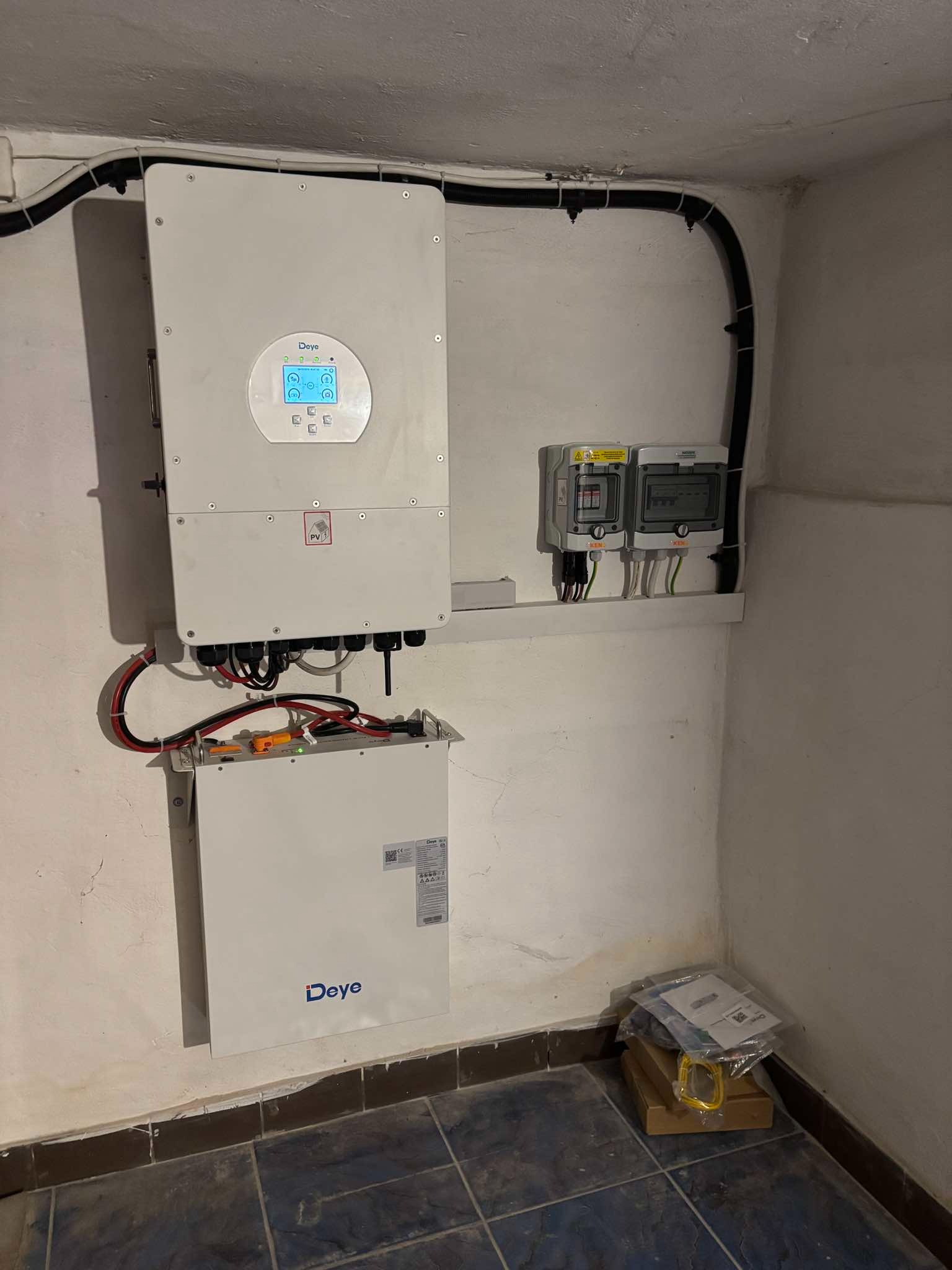 Zamontowany falownik i bateria Deye w pomieszczeniu. Widoczne zabezpieczenia elektryczne i okablowanie. Całość w surowym wnętrzu z ciemną podłogą.