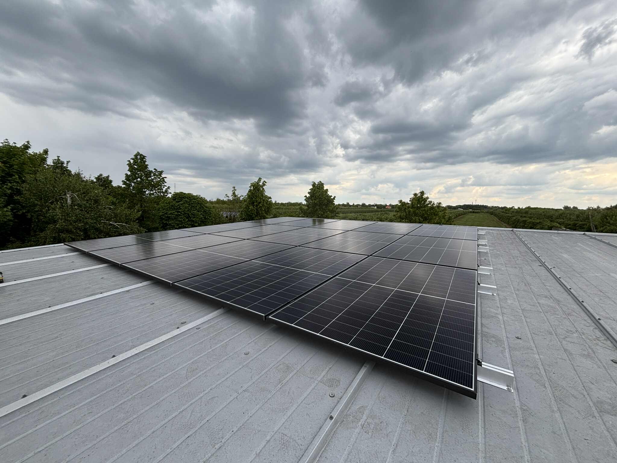 Panel słoneczny na szarym dachu, widok z dołu. Na horyzoncie zieleń drzew, pochmurne niebo. Widoczne ślady montażu.