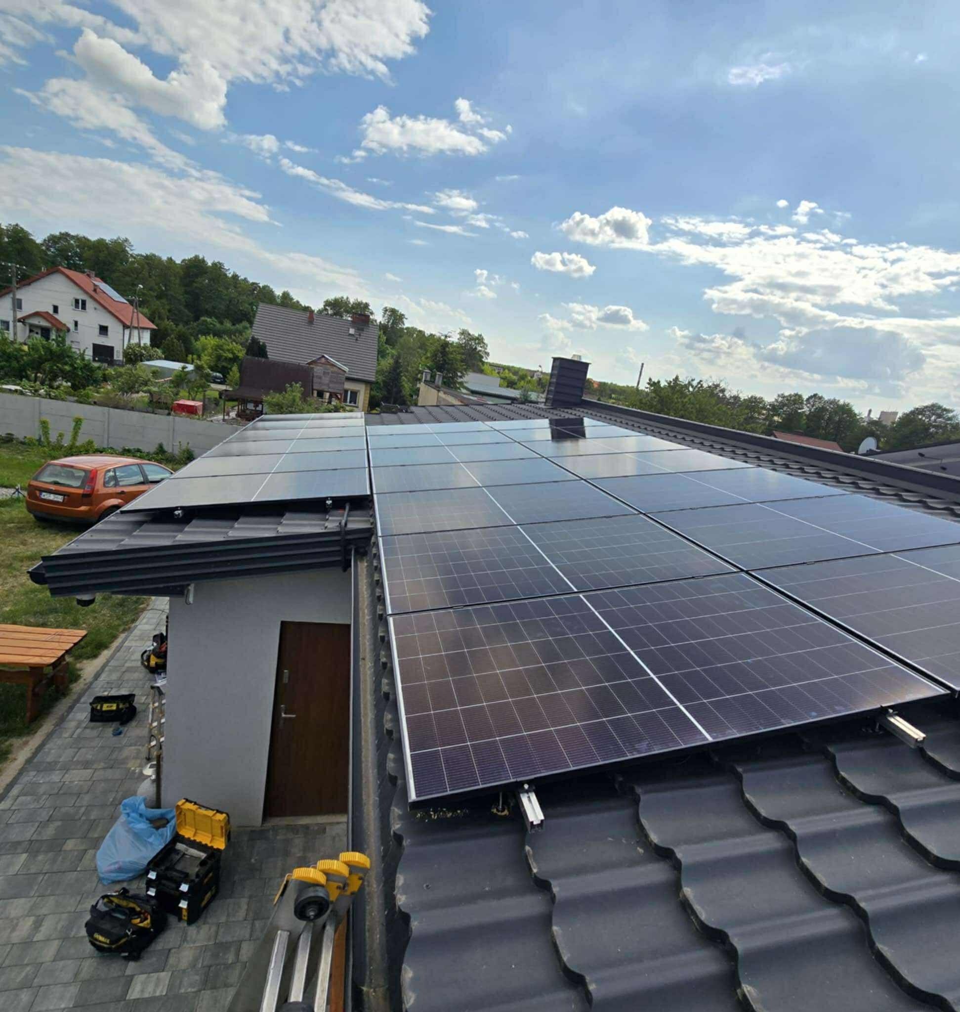 Panele fotowoltaiczne zamontowane na dachu domu jednorodzinnego, widoczne narzędzia pracy na chodniku obok budynku. Słoneczny dzień.