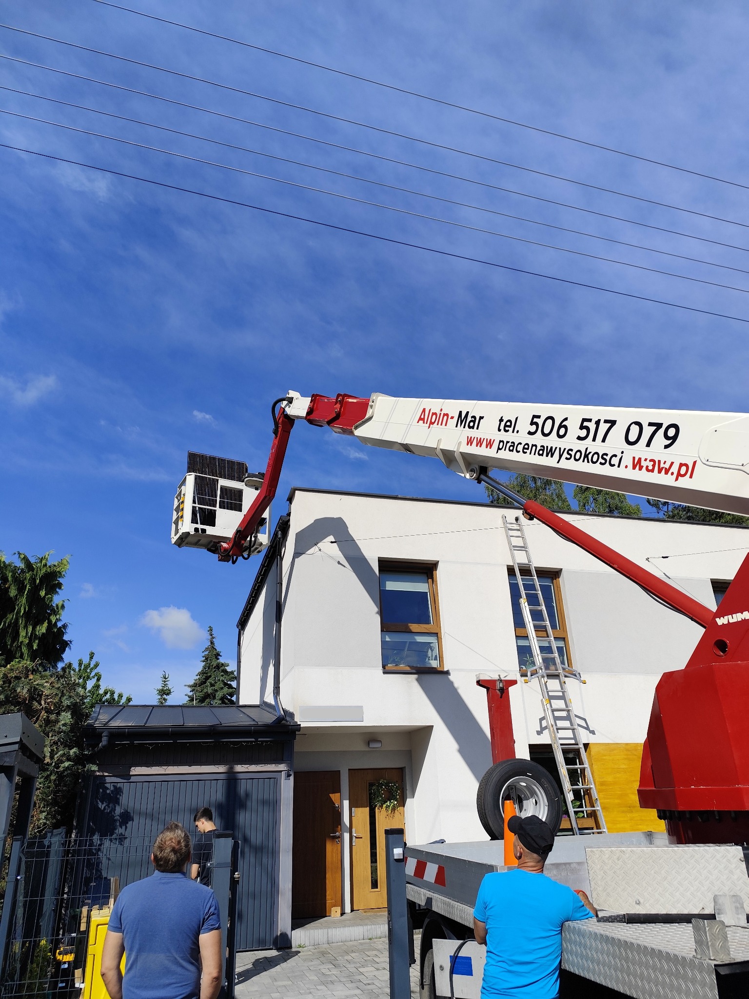 Montaż paneli fotowoltaicznych na dachu domu jednorodzinnego z użyciem podnośnika koszowego firmy Alpin-Mar. Widoczni pracownicy i drabina oparta o ścianę budynku.