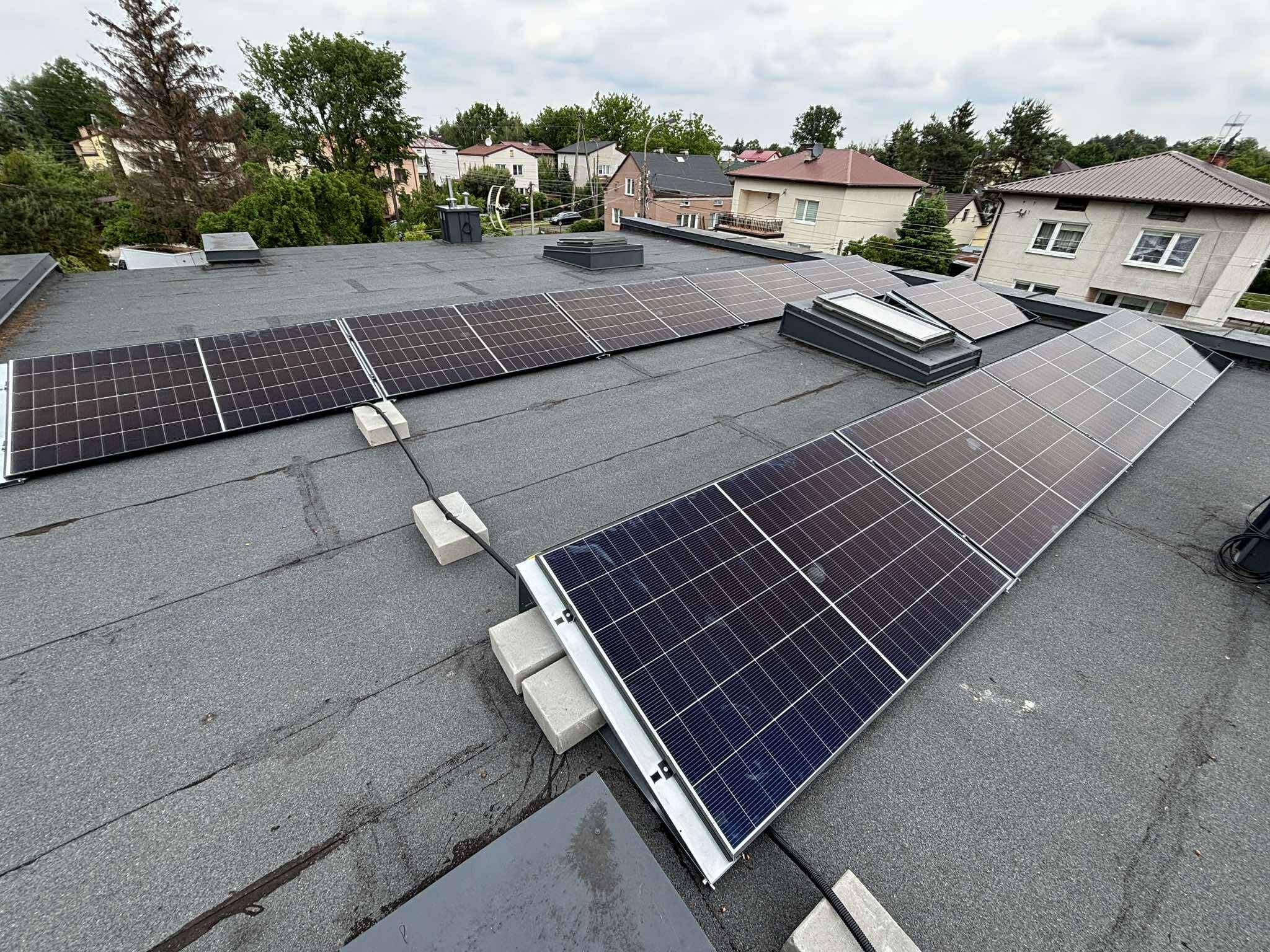Instalacja paneli słonecznych na płaskim dachu budynku. Panele ustawione na betonowych blokach. Widok na domy i drzewa w tle.
