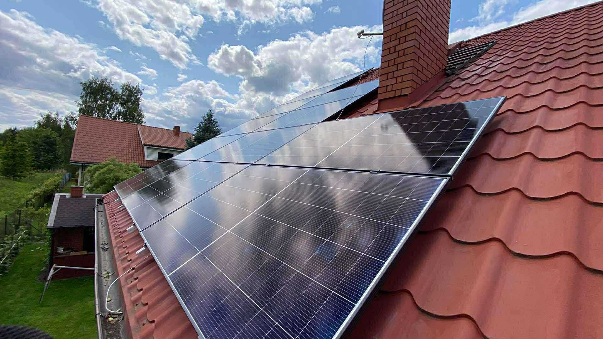 Panele słoneczne na czerwonym dachu, komin z cegły w tle, błękitne niebo z chmurami. Widok z dołu, instalacja fotowoltaiczna na domu jednorodzinnym.