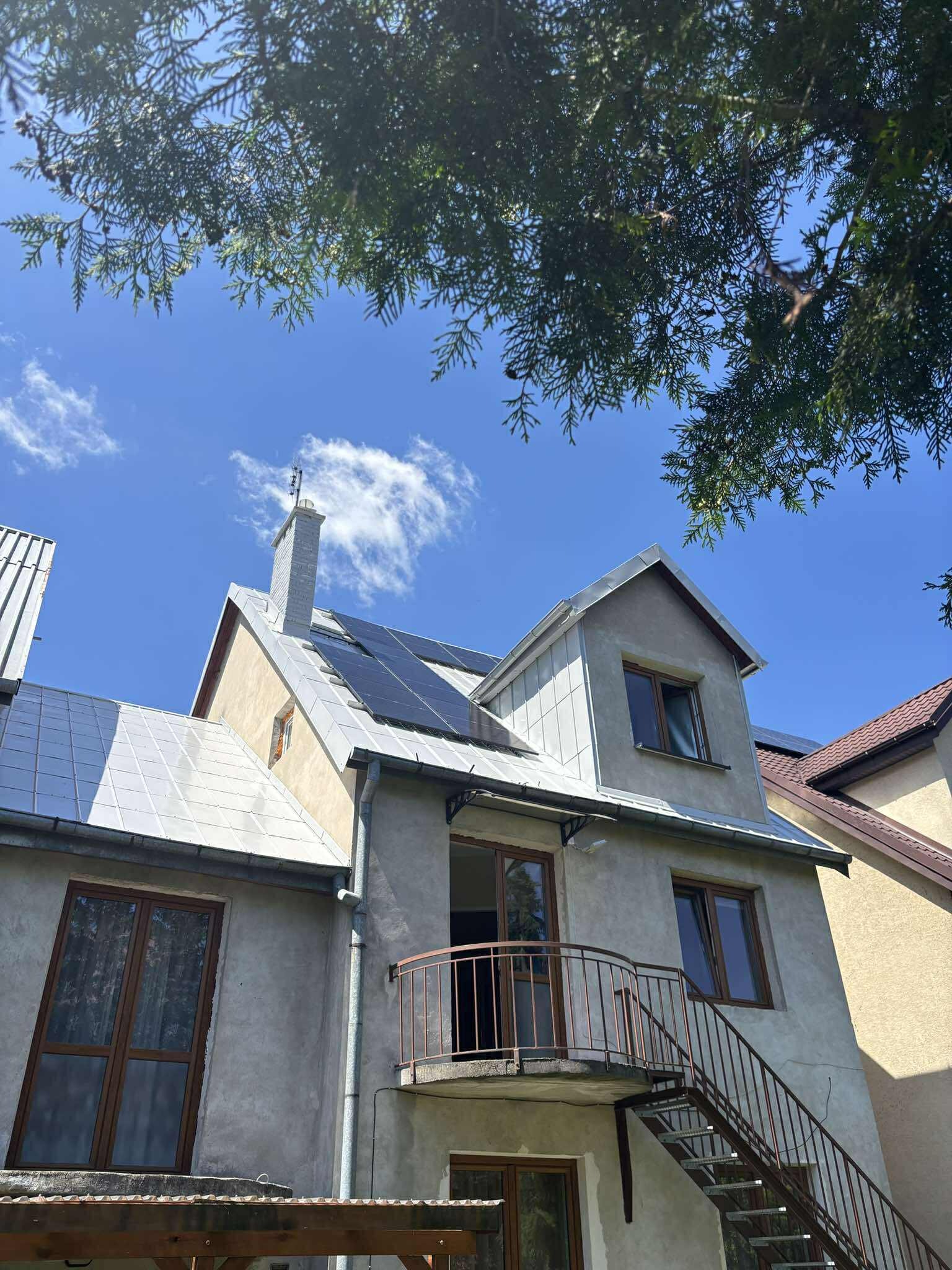 Panele słoneczne na dachu domu jednorodzinnego w Żyrardowie, widziane z dołu, z fragmentem korony drzewa w rogu. Błękitne niebo w tle.