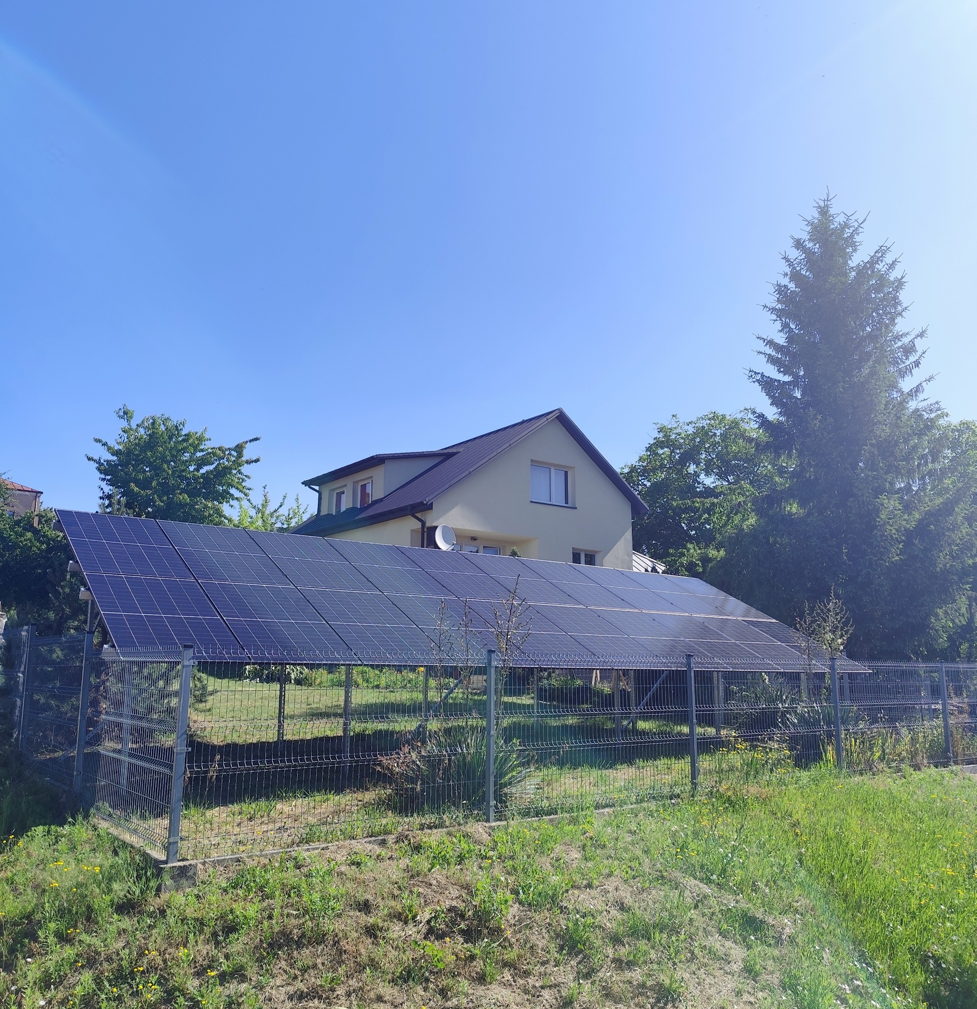 Rząd paneli fotowoltaicznych na metalowej konstrukcji, z domem w tle, otoczony zielenią w słoneczny dzień. Widok z poziomu gruntu.