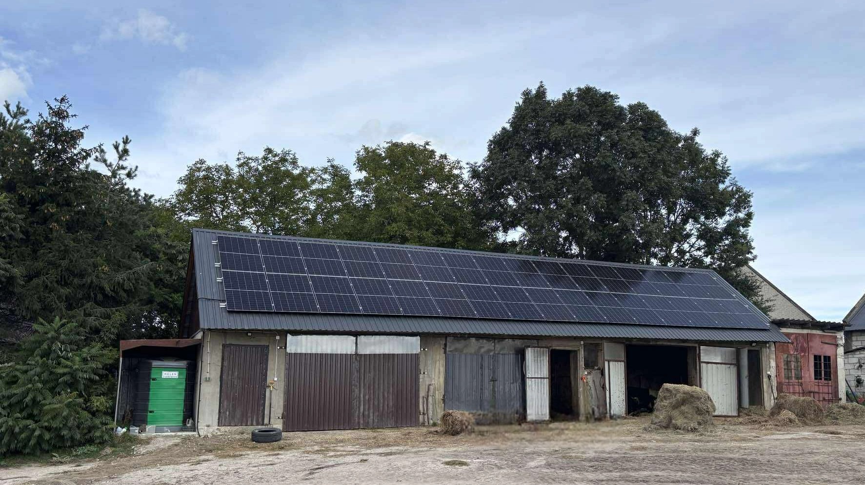 Instalacja paneli słonecznych na dachu budynku gospodarczego z szarej blachy falistej, otoczonego drzewami i zieloną toaletą przenośną.