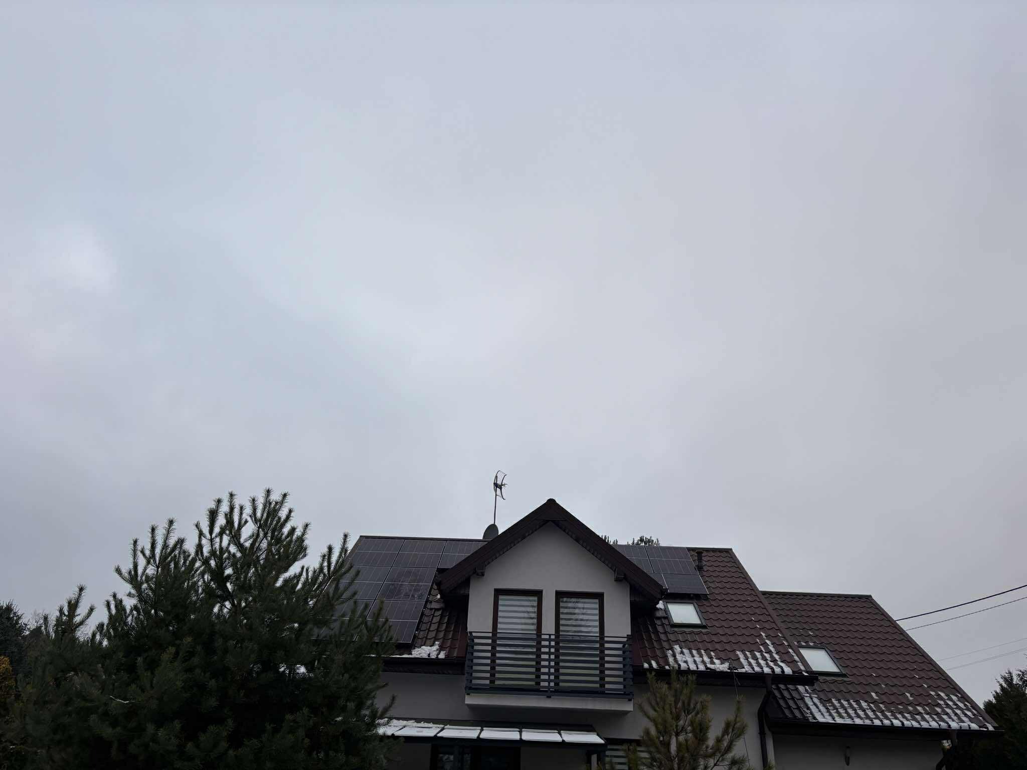 Dom z panelami słonecznymi na dachu w pochmurny dzień. Widoczne panele fotowoltaiczne, balkon i antena. Częściowo ośnieżony dach z dachówką.