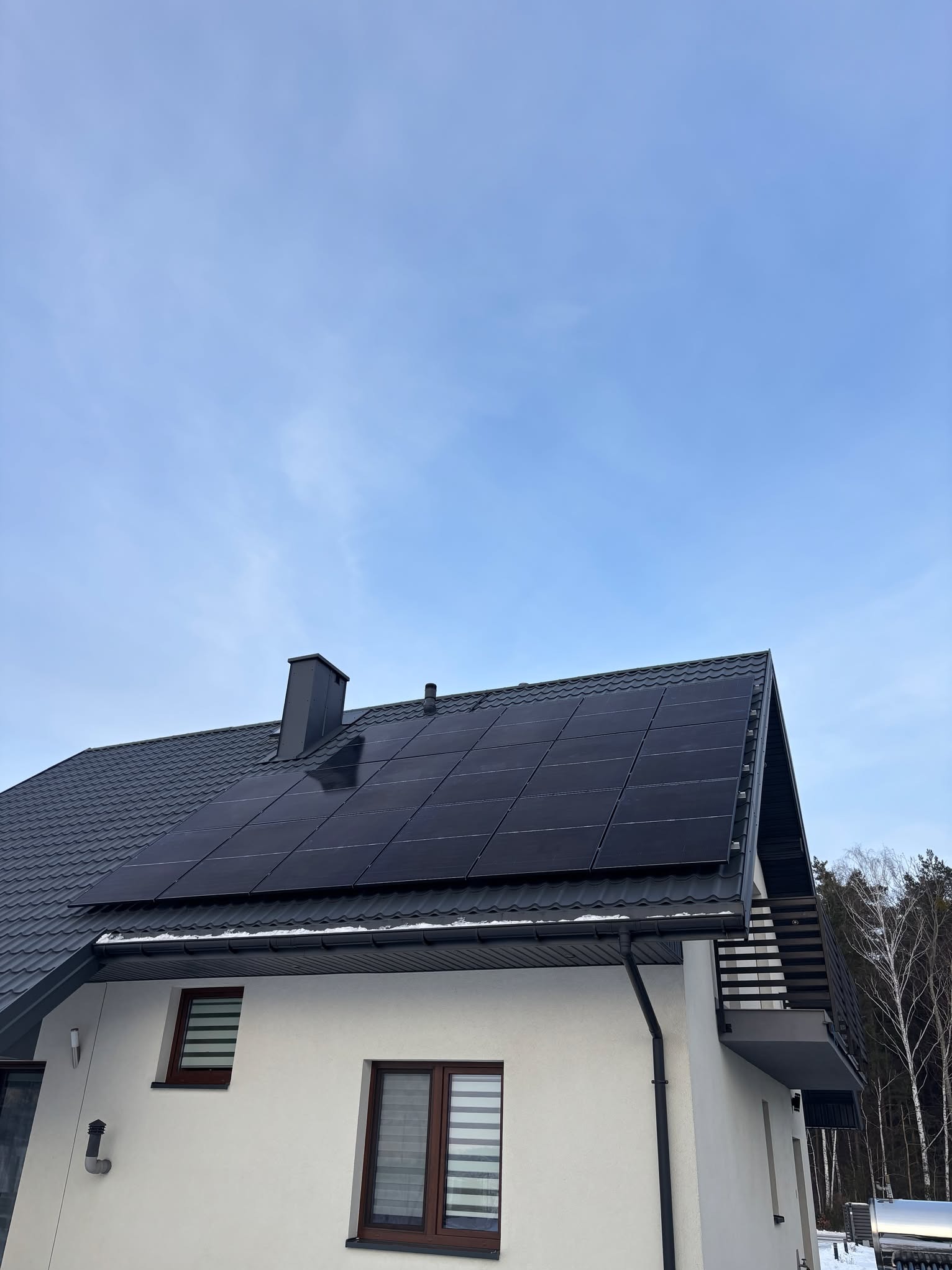 Czarny panel fotowoltaiczny na dachu domu z szarej blachy dachówkowej, widok z dołu na tle błękitnego nieba i drzew. Część elewacji w kolorze białym.