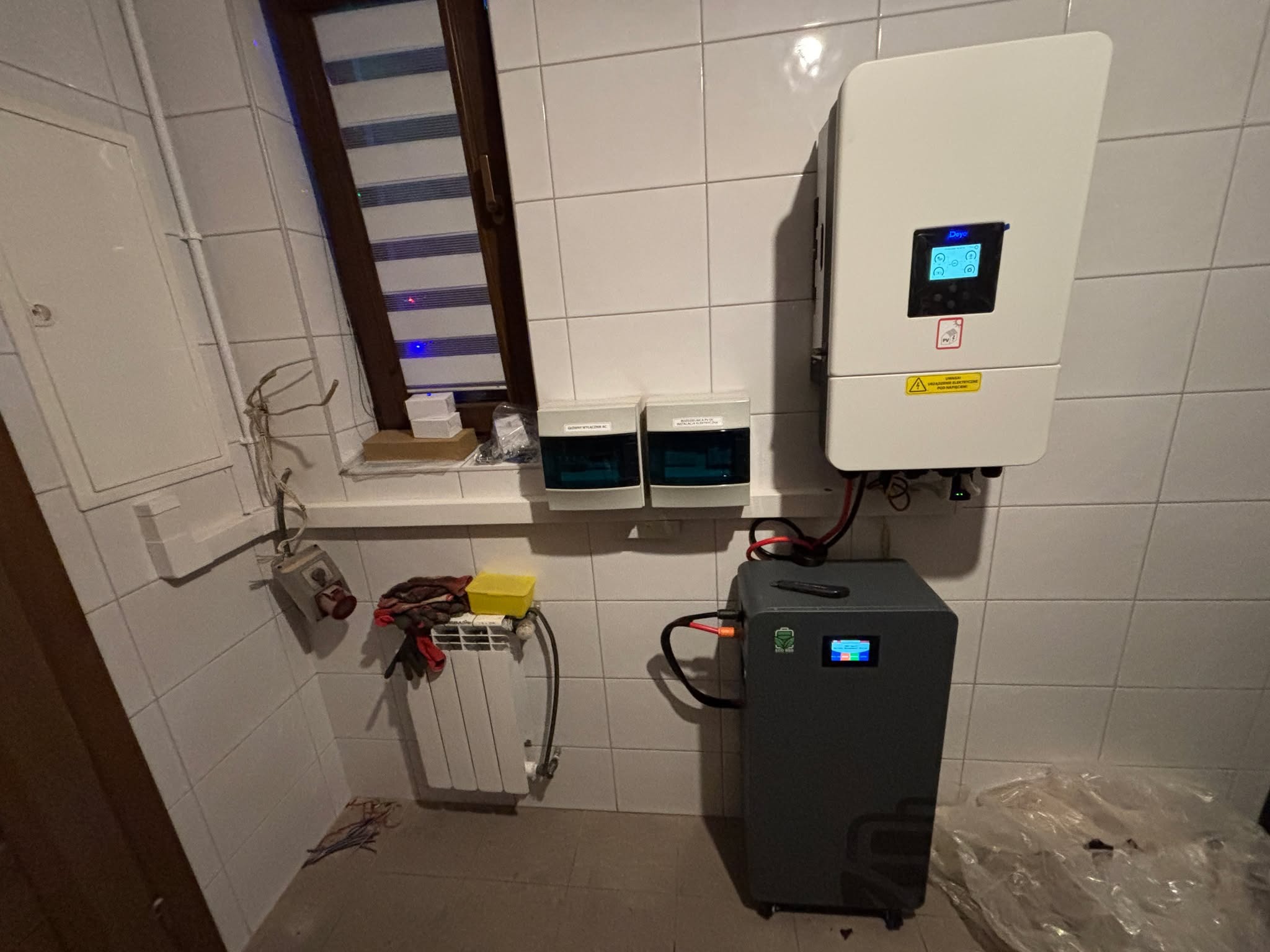 Instalacja magazynu energii w pomieszczeniu z wykończeniem w płytkach, Żyrardów. Widoczny inwerter, akumulator i zabezpieczenia. Elementy instalacji elektrycznej.