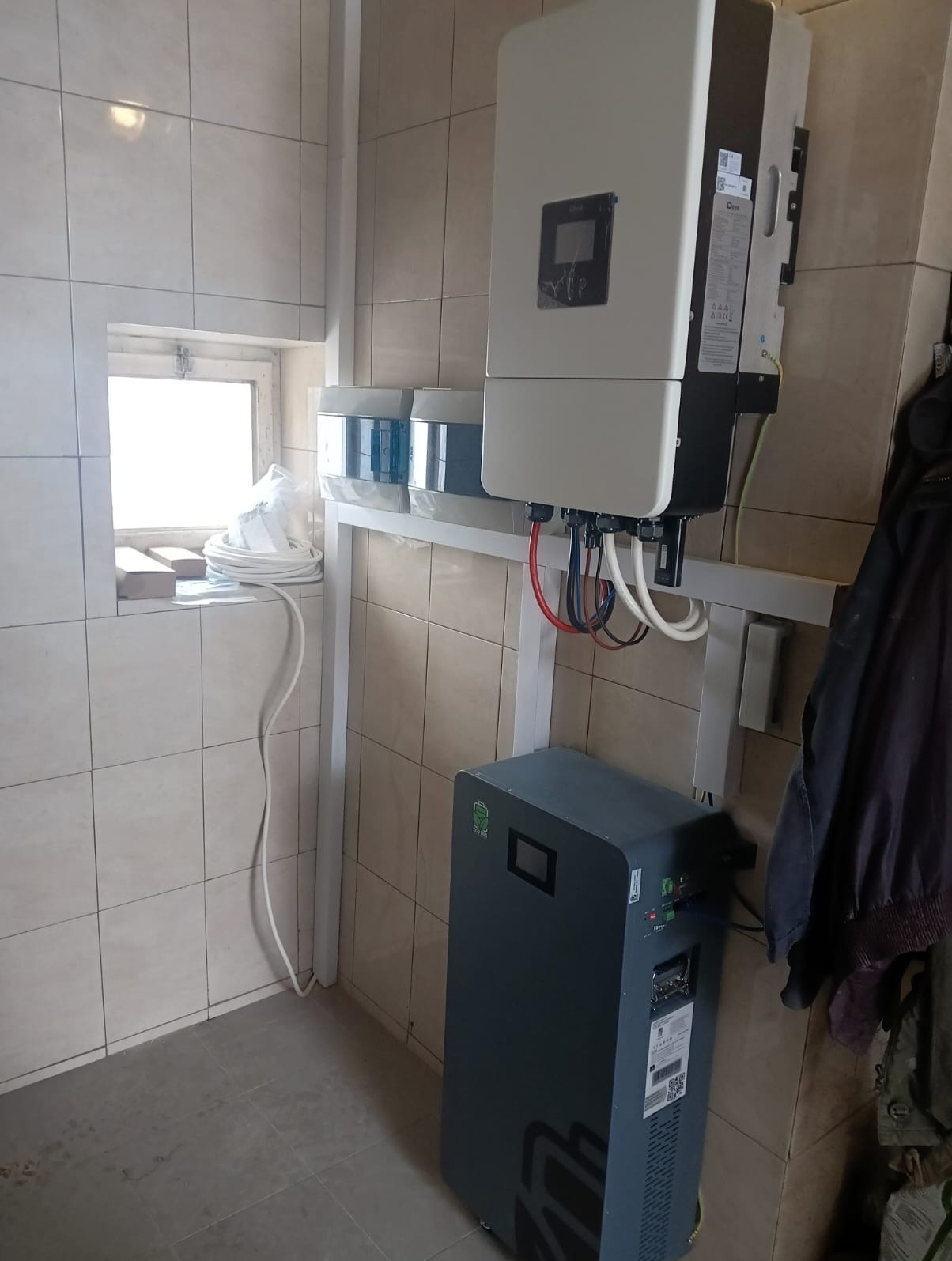 Zamontowany inwerter i magazyn energii w pomieszczeniu z kafelkami. Widoczne kable i przewody. Elementy instalacji przymocowane do ściany na metalowym stelażu.