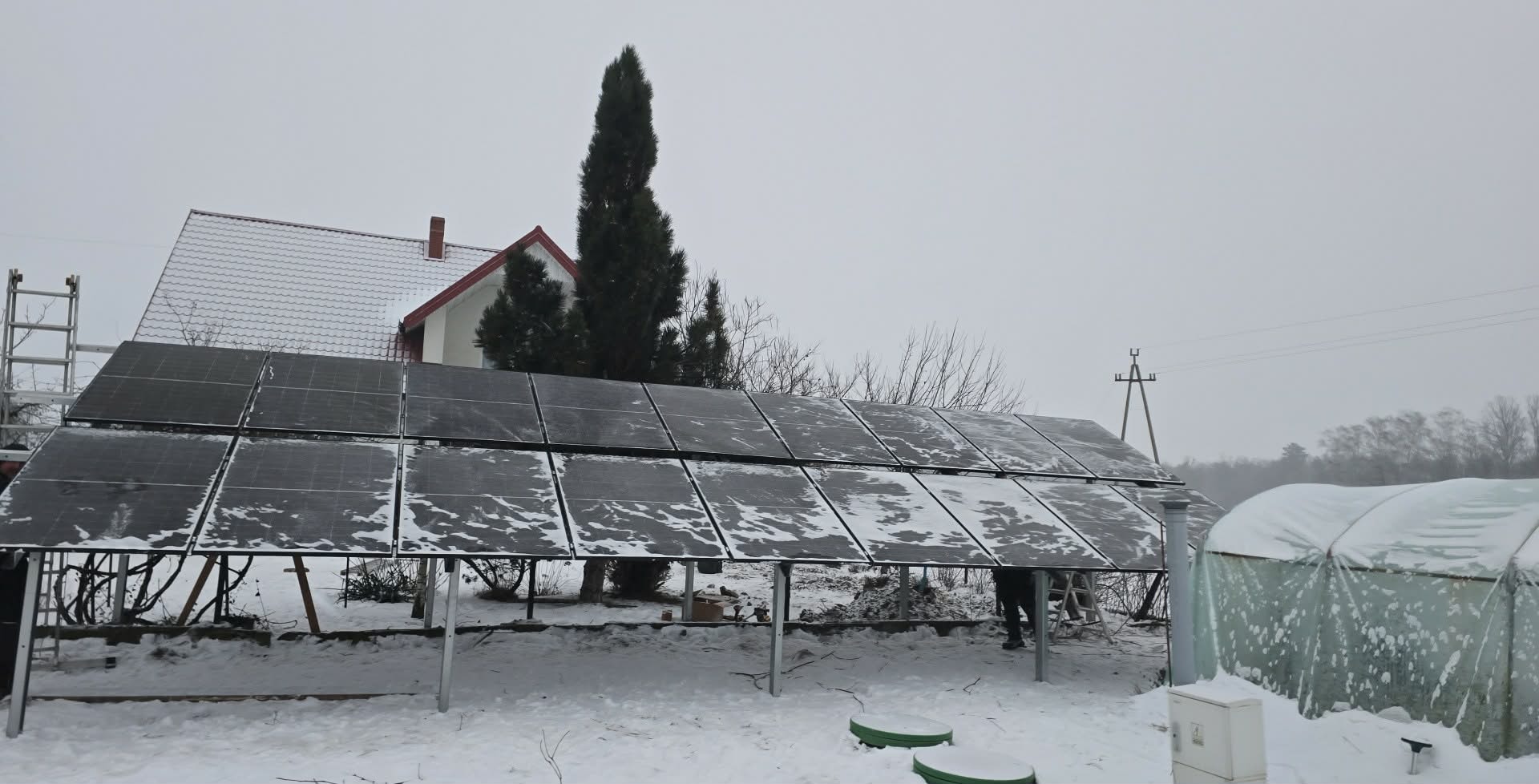 Zimowy montaż paneli słonecznych na konstrukcji wsporczej, z domem w tle. Panele pokryte śniegiem, pochmurne niebo. Widoczna drabina i częściowo zadaszona szklarnia.