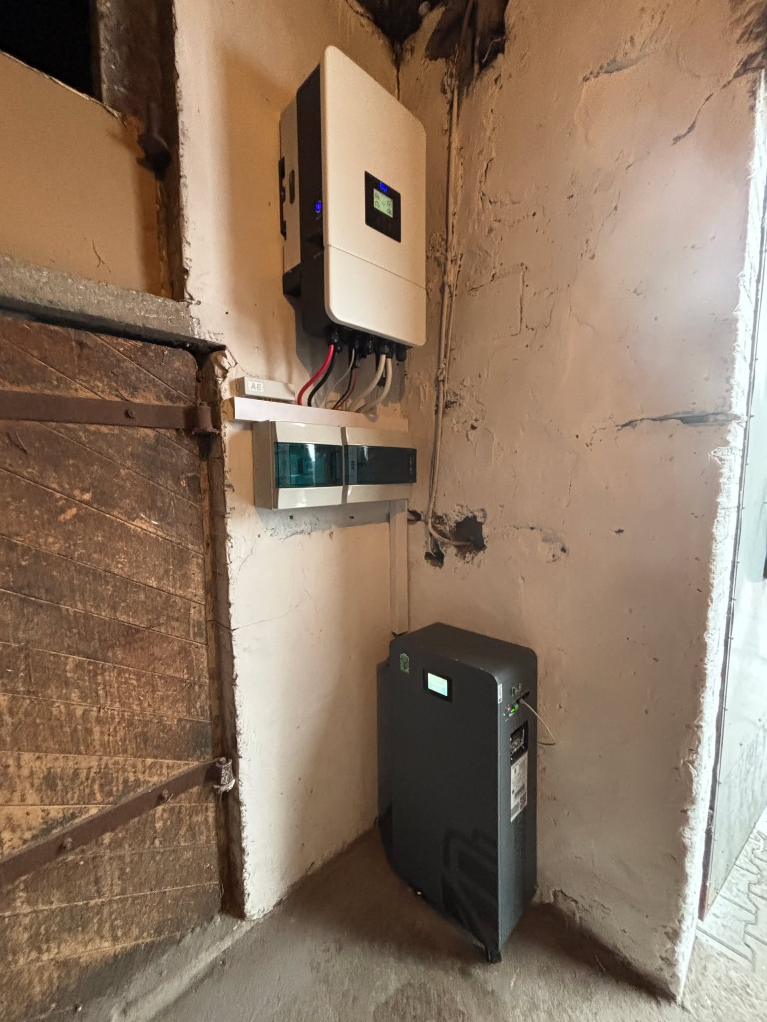Instalacja wewnątrz budynku: biały inwerter, szara skrzynka rozdzielcza i czarny magazyn energii na tle ściany z surowym tynkiem i fragmentem drewnianych drzwi.
