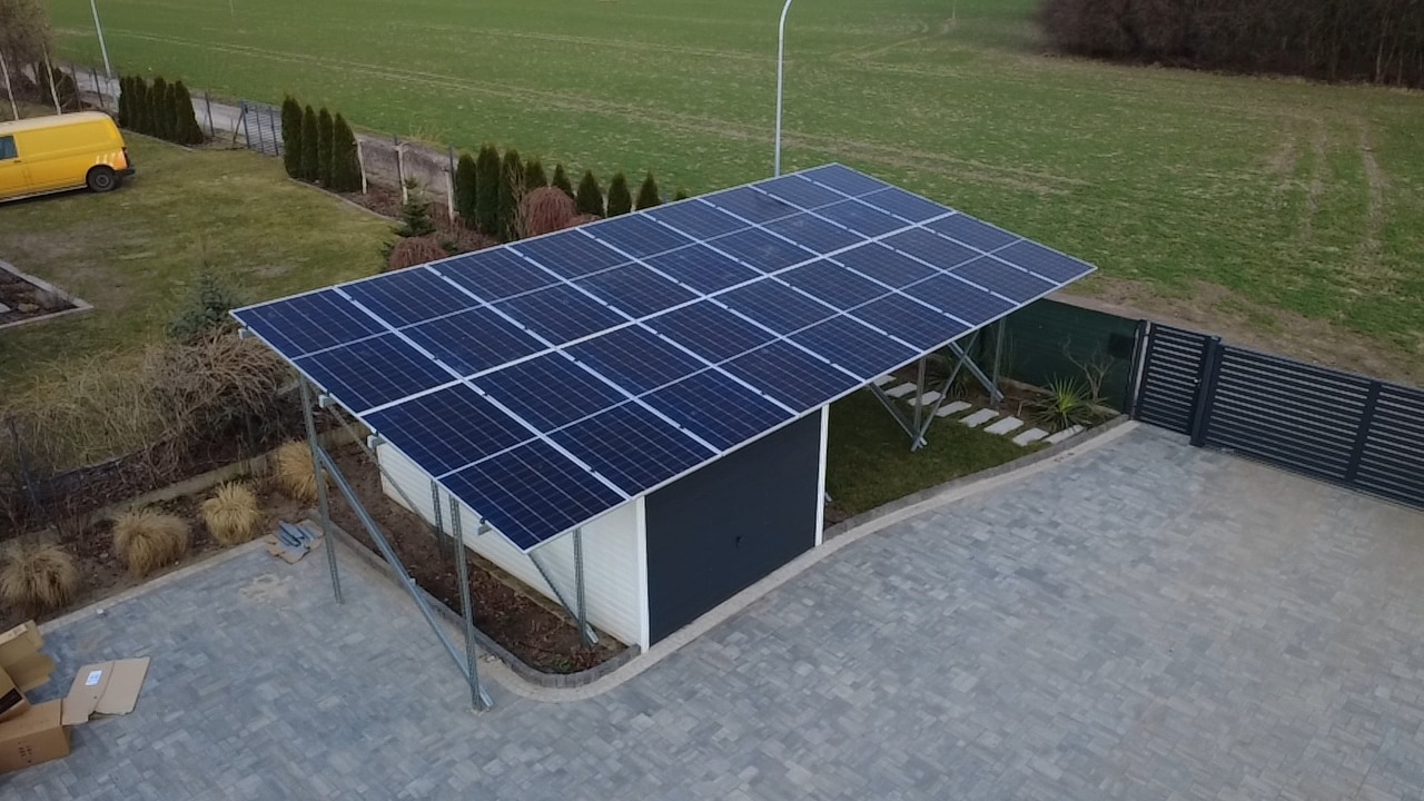 Zadaszenie z paneli słonecznych nad garażem, widok z góry. Na podwórku kostka brukowa, trawnik i żółty samochód dostawczy w tle. Nowoczesna instalacja.