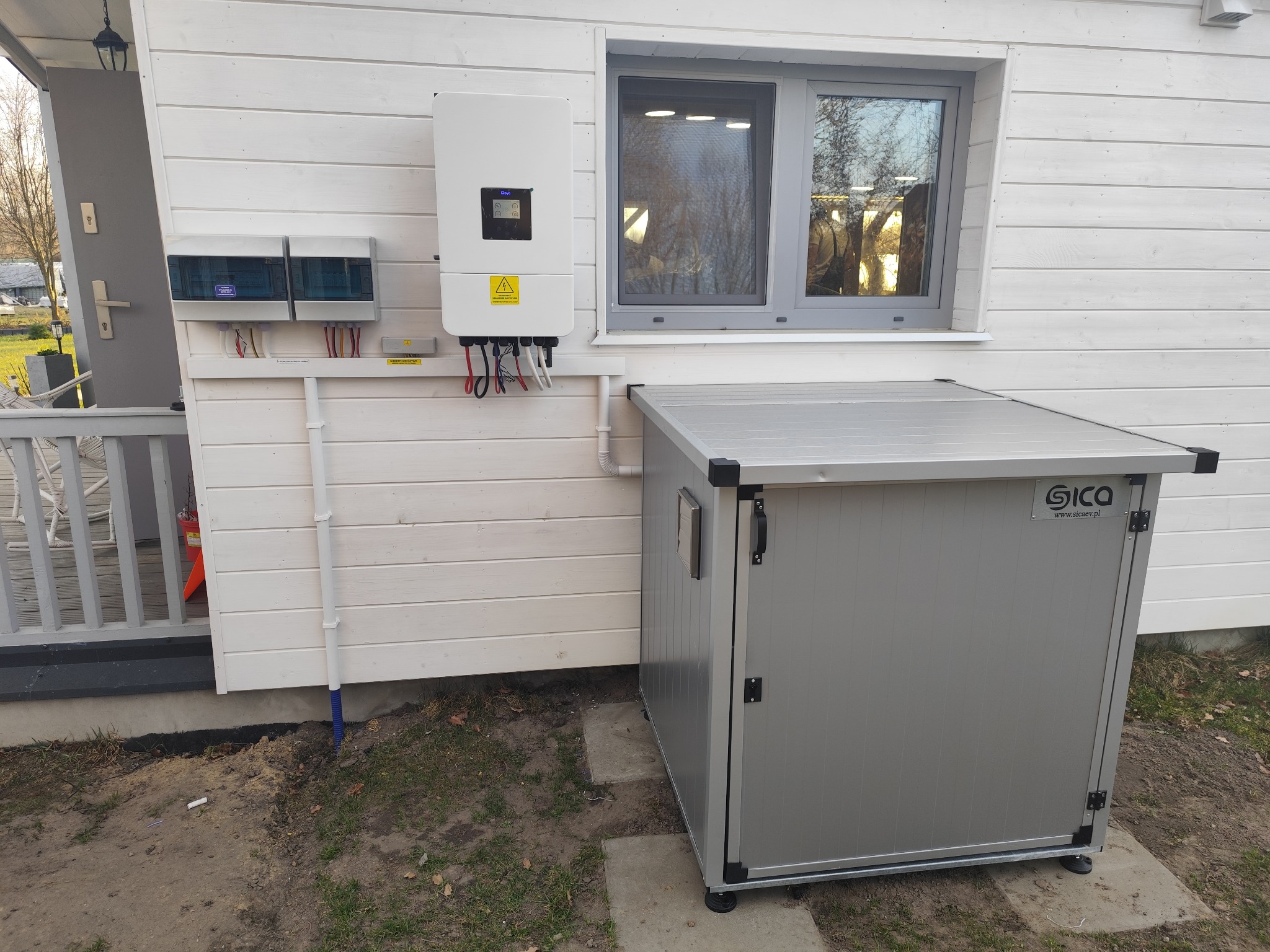 Biała skrzynka z instalacją elektryczną i szarym magazynem na zewnątrz domu z drewnianą elewacją. Widoczne okno i elementy instalacji.