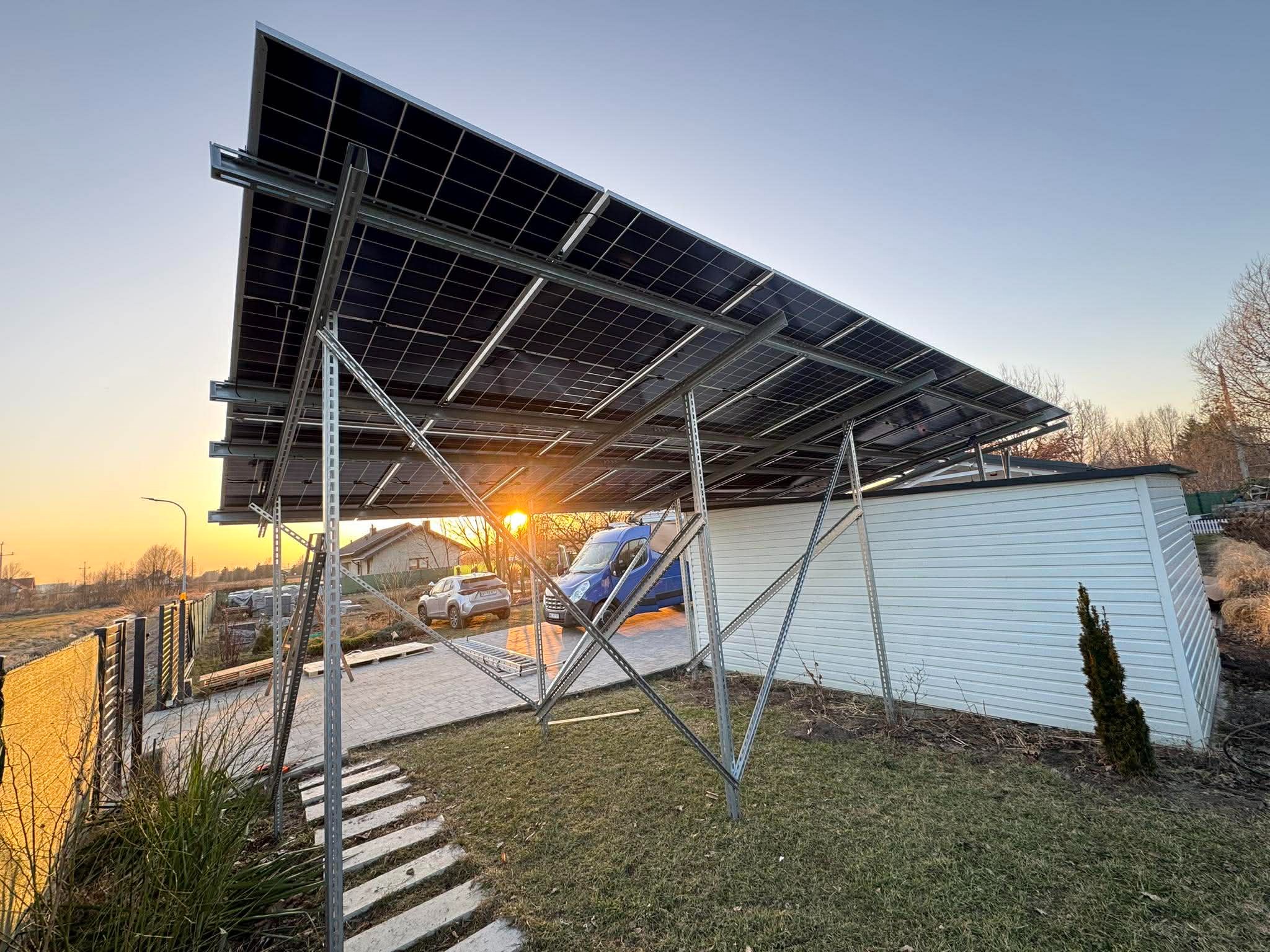 Montaż paneli fotowoltaicznych na konstrukcji wsporczej, widok z dołu na tle zachodzącego słońca. W tle budynek i samochody.
