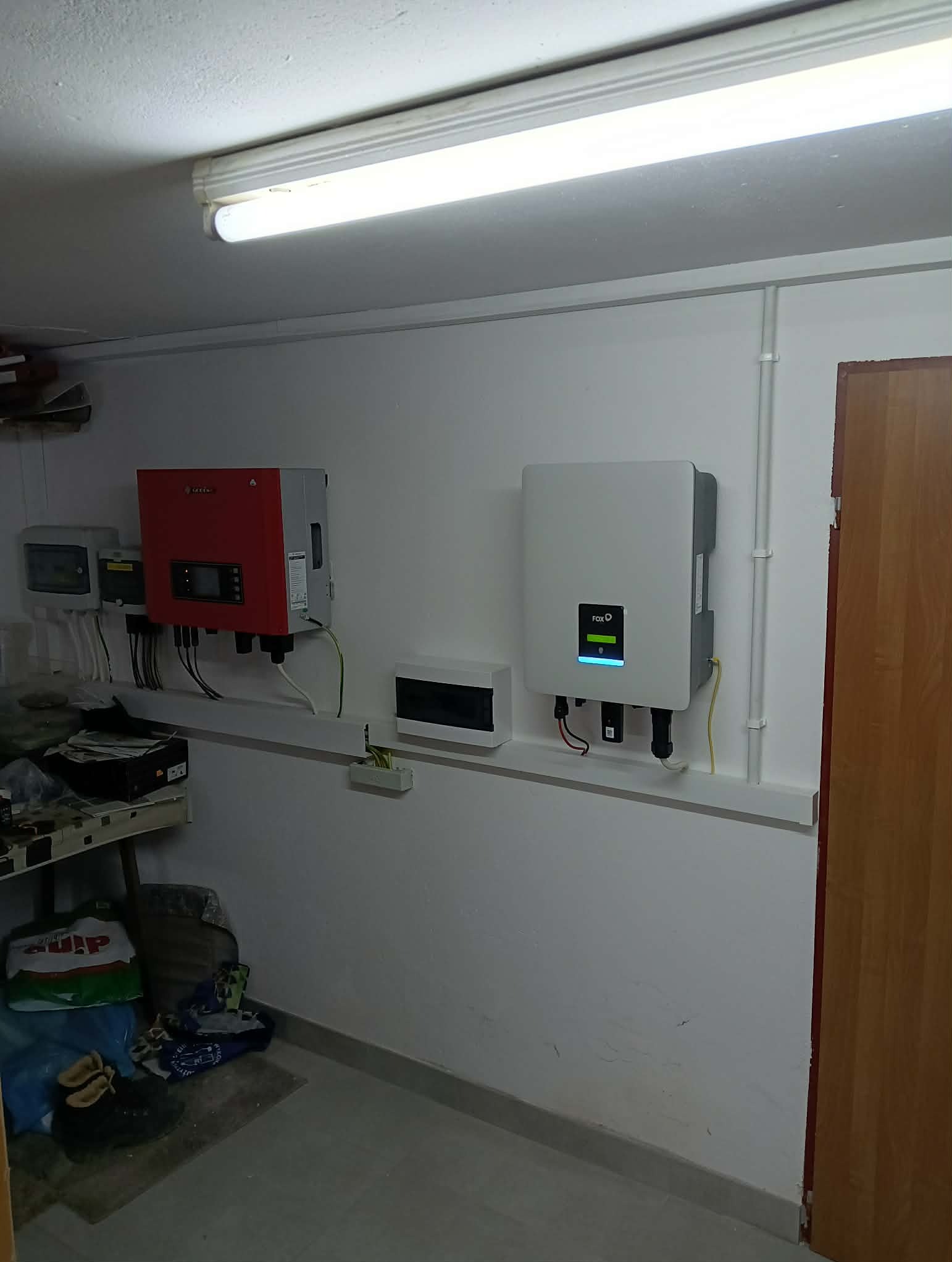 Dwa inwertery, czerwony i biały, zamontowane na ścianie w pomieszczeniu. Widoczne kable i elementy instalacji elektrycznej. Górne oświetlenie jarzeniowe.