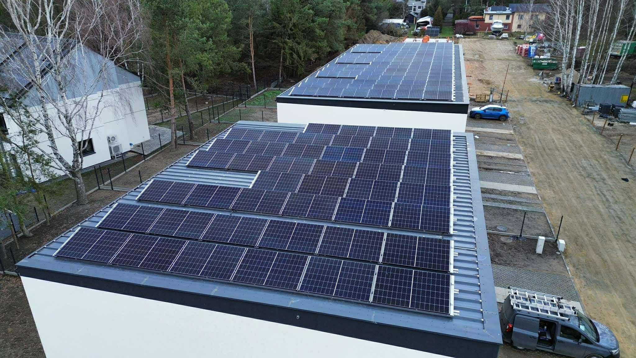 Dwa budynki z panelami słonecznymi na dachu, widok z góry. Na drugim planie las i zabudowa mieszkalna. Na dachu jednego z budynków brakuje kilku paneli.