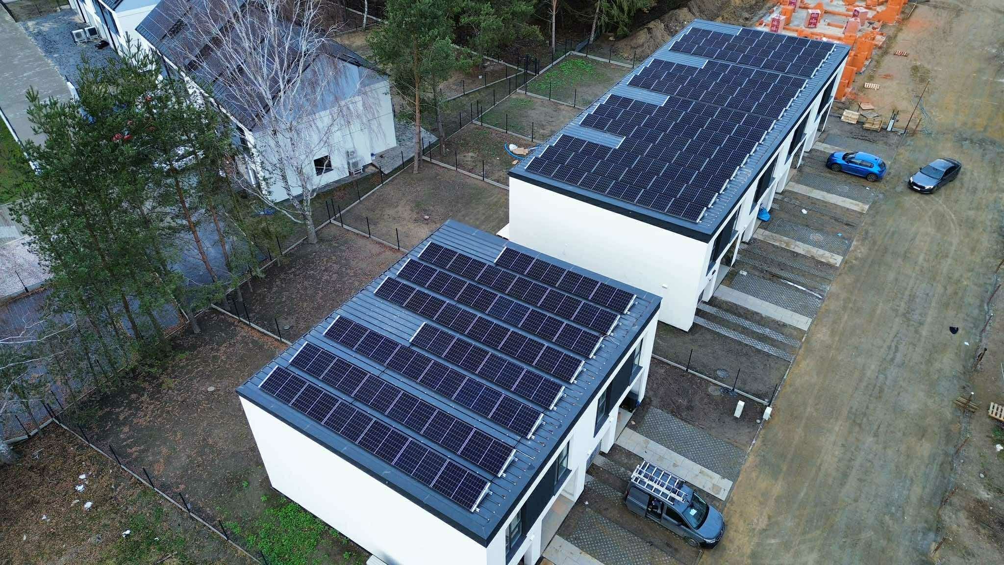 Widok z góry na dwa nowoczesne budynki z panelami słonecznymi na dachach, zaparkowane samochody i otaczającą zieleń. Jeden budynek w trakcie budowy w tle.