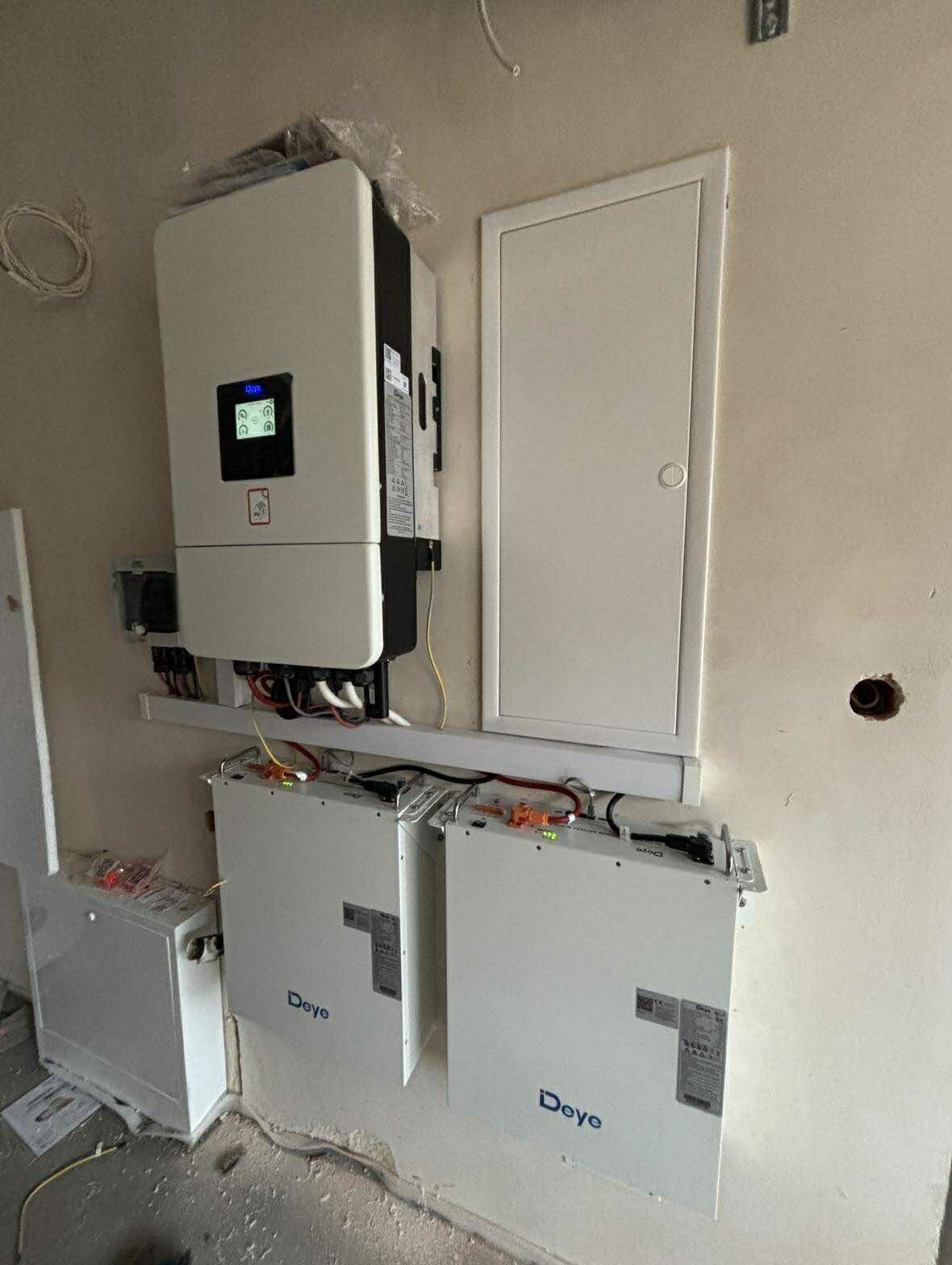 Zamontowany inwerter i dwa akumulatory Deye na ścianie wewnątrz budynku. Widoczne okablowanie i elementy instalacji. Biała ściana z widocznymi niedoskonałościami.