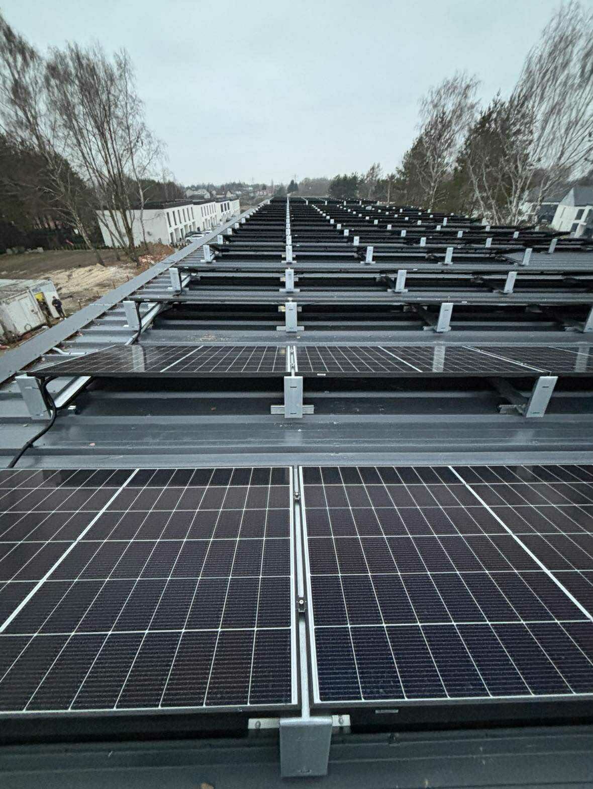 Rząd paneli fotowoltaicznych na szarym dachu budynku, zamontowanych na metalowych wspornikach. W tle drzewa i budynki w pochmurny dzień.