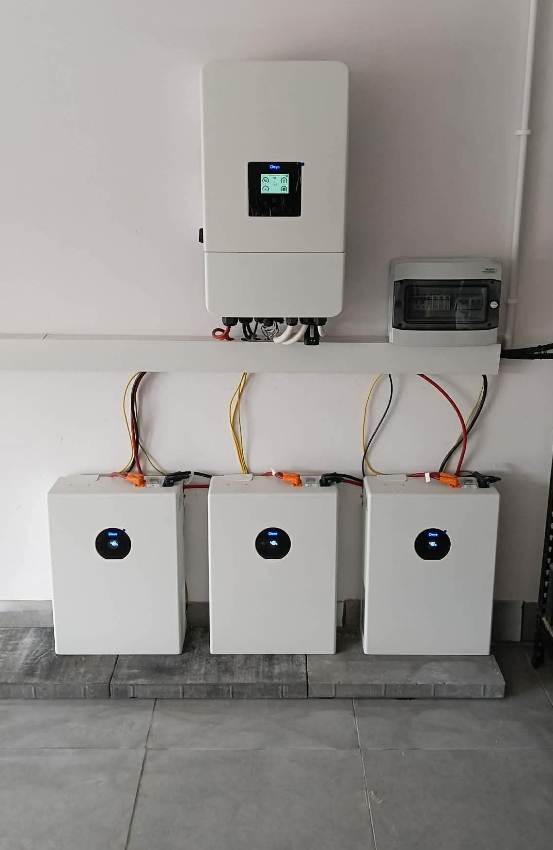Trzy białe moduły magazynu energii Deye, zamontowane na szarej podłodze, z podłączonymi kablami i inwerterem na ścianie. Widoczna skrzynka elektryczna.