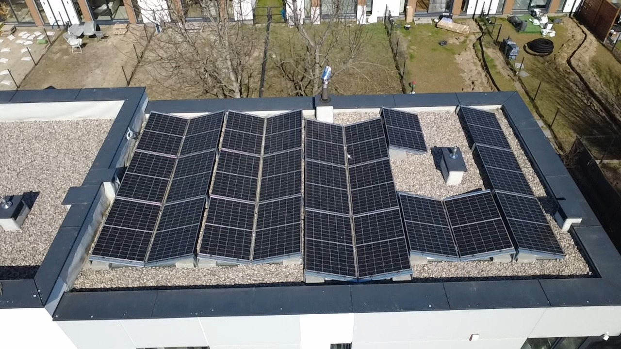 Instalacja paneli słonecznych na płaskim dachu budynku mieszkalnego, widok z góry. Panele ułożone w zygzak na żwirowym podłożu. W tle drzewa i zabudowa.