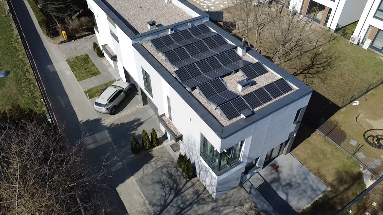Nowoczesny dom z panelami słonecznymi na dachu, widok z góry. Biała elewacja, szary dach i zadbany ogród. Samochód zaparkowany przed budynkiem.