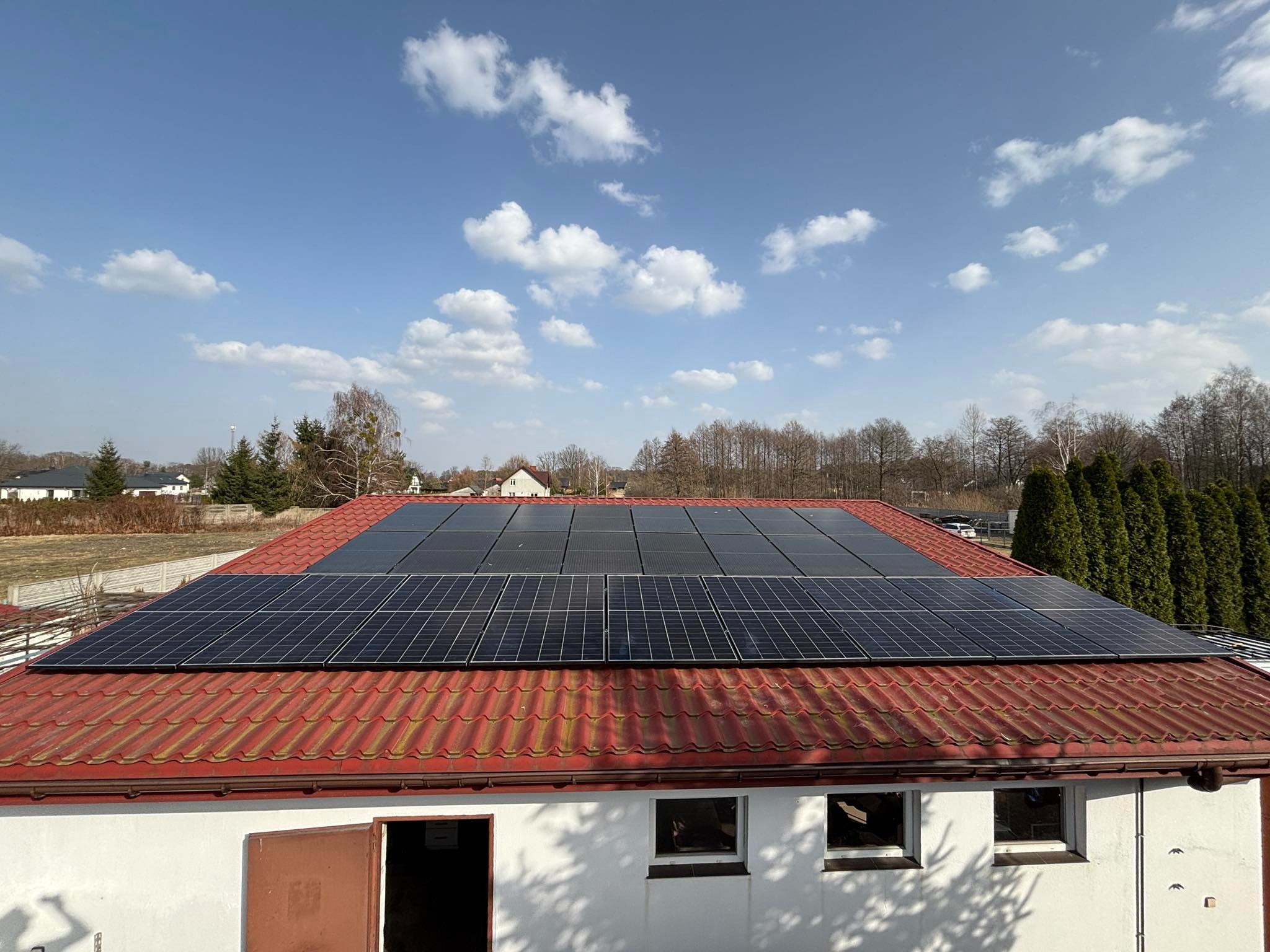 Panele słoneczne na czerwonym dachu budynku z białymi ścianami, błękitne niebo z chmurami w tle. Widok z góry, lekko z boku. Instalacja fotowoltaiczna.