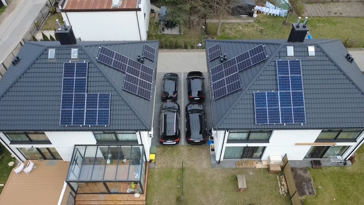 Dwa domy bliźniacze z panelami słonecznymi na dachach, cztery czarne samochody zaparkowane pomiędzy budynkami, widok z lotu ptaka.